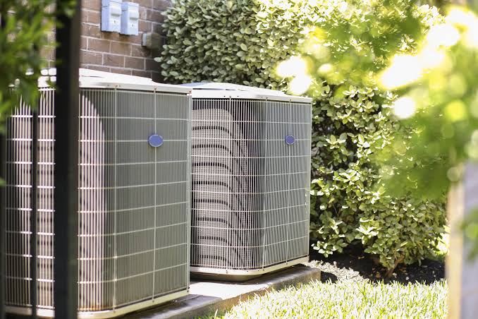 Two residential AC condenser units beside a home with greenery behind